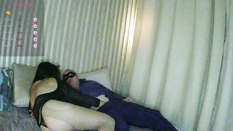 A candid video shows a dark-haired woman in a black lace bodysuit and a blindfold kissing a bald man in a purple shirt and dark pants on a bed with beige curtains.