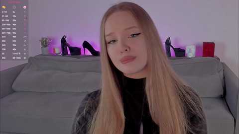 Video of a young, fair-skinned woman with long, straight blonde hair, wearing a black top, sitting on a grey couch. Behind her, high heels and candles on a white wall.