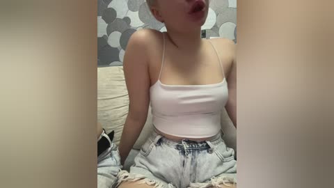 Video of a fair-skinned woman with a short bob haircut, wearing a white crop top and high-waisted light blue jeans. She's seated on a beige couch with a gray and white patterned wall behind her.