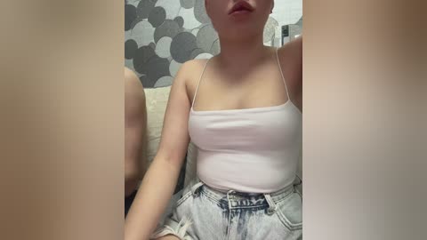 Video of a woman with fair skin, light hair, wearing a white spaghetti-strap tank top and high-waisted light-wash denim shorts, seated indoors with gray patterned wallpaper background.