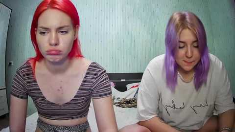 Media: Video of two young women with vivid red and purple hair, wearing striped and plain white shirts, sitting on a bed in a room with green walls.