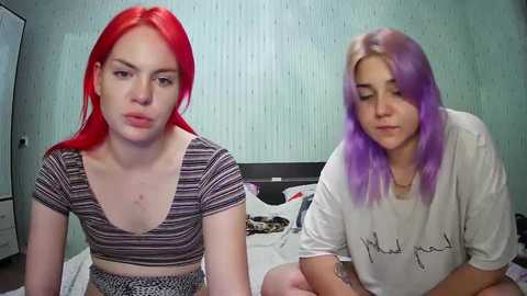 Media: Video of two young women with vivid red and purple hair, wearing striped crop tops, sitting on a bed in a room with light blue, textured wallpaper.