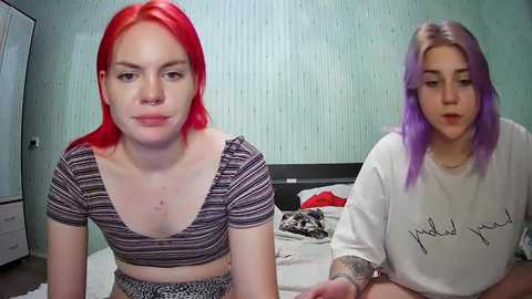 Media: Video of two women with vibrant red and lavender hair, sitting on a bed with a striped crop top and a white T-shirt, respectively.