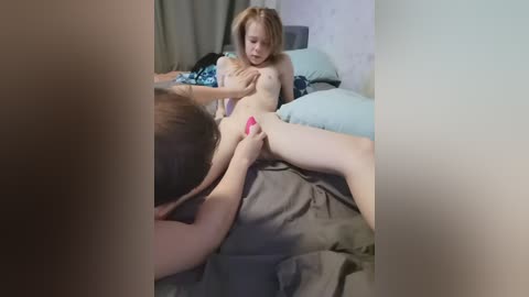 Media: Video of a naked young woman with long red hair lying on a bed, being pleasured by a man with short brown hair; both are Caucasian, in a bedroom with light-colored walls.