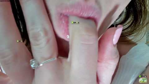 Video of a close-up of a woman with fair skin, medium brown hair, and a pink manicure, using her fingers to hold a small, shiny object in her mouth.