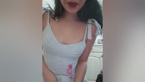 Media: Video of a woman with long black hair, wearing a light blue, sleeveless dress with pink floral embroidery, seated in a kitchen with white cabinets and a black purse.