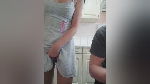 Media: Video of a slender, light-skinned woman in a light blue tank top and shorts, standing in a kitchen with white cabinets and beige countertops. A man in a dark t-shirt is partially visible.