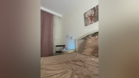 Media: Video of a minimalist, beige-colored bedroom with a neatly made bed, a white headboard, a small nightstand, and a decorative wall painting.