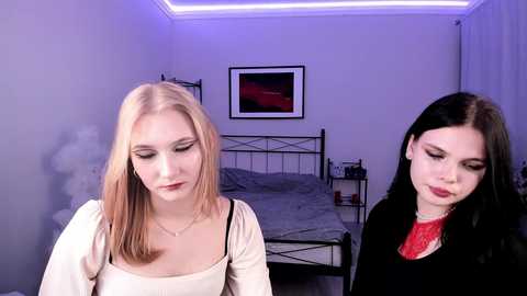 Video of two young women, one blonde in a beige top, the other brunette in a red dress, sitting in a dimly lit bedroom with a bed, framed painting, and blue light.