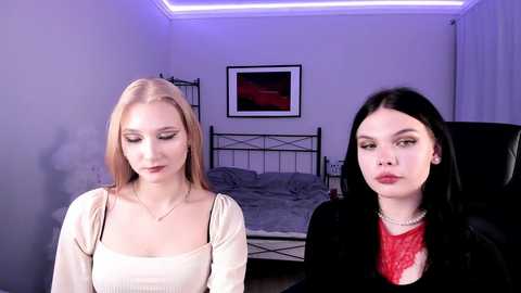 Video of two women: a blonde with pale skin in a beige top and a brunette with fair skin in a red top, sitting in a dimly lit room with a bed and framed art in the background.