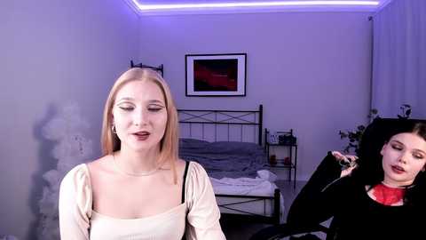Video of a pale-skinned blonde woman in a beige top, standing next to a brunette woman with a black hat, in a dimly-lit, modern bedroom with gray walls, a bed, and framed art.