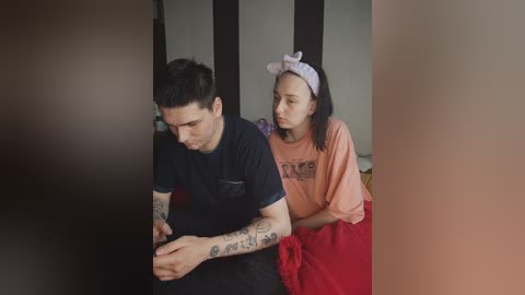 Video of a tattooed young man with short dark hair, wearing a black T-shirt, sitting next to a young woman with straight black hair and a lavender headband, wearing an orange T-shirt, in a dimly lit room.