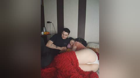 Video of a man with tattoos, wearing a black shirt, leaning over a woman with fair skin, wearing a red fuzzy blanket, lying on a bed with a white pillow.