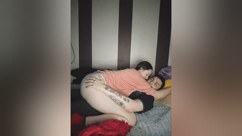 A video shows a woman with a tattooed leg embracing a man on a bed with red and gray blankets, set against a dark wooden backdrop.