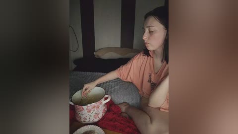 Video of a young Asian woman with long black hair, wearing an orange shirt, sitting on a bed with a red blanket. She is pouring milk from a pink floral cup into a white bowl.