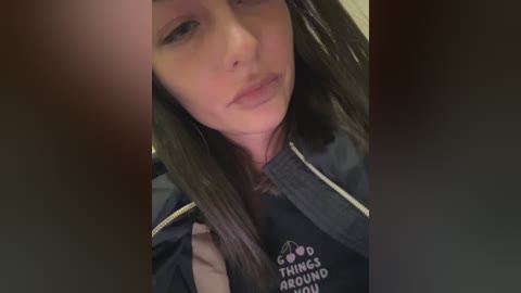 Media: Video of a young woman with fair skin, long brown hair, and a pouty expression, wearing a black hoodie with \"GOOD THINGS AROUND US\" text, taken indoors.