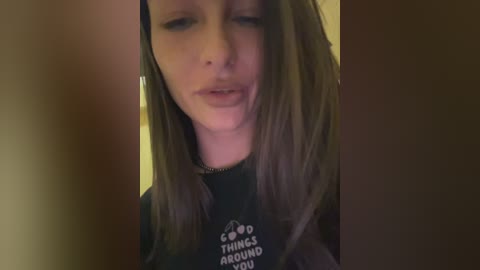 Media: A video of a young woman with long, straight brown hair, wearing a black t-shirt with a white logo, looking down. The background is blurred.