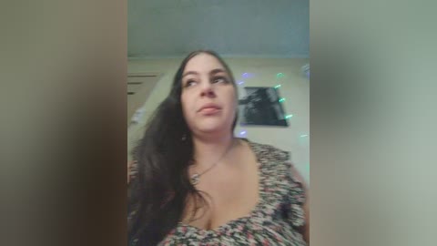 A video of a smiling, light-skinned woman with long, wavy black hair, wearing a floral-patterned blouse, standing in a dimly lit room with string lights and a framed photo on the wall.