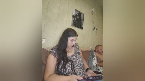 Video of a young woman with long black hair, wearing a floral dress, sitting on a brown sofa, reading a book, in a dimly lit, beige-walled room with a black-and-white photo and fairy lights on the wall.