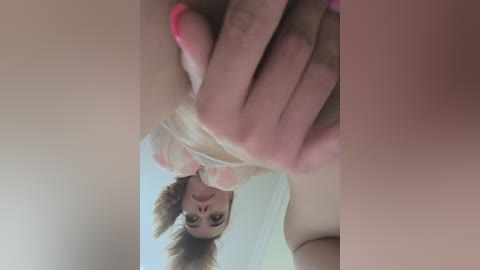 Media: Video of a fair-skinned woman with brown hair and pink nails, wearing a light-colored shirt, seen from a low angle. She holds her hand up, partially covering her face, and the background is blurred.