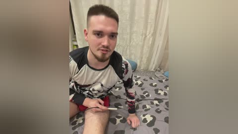 Media: Video of a young man with short brown hair, wearing a camo-patterned long-sleeve shirt and red shorts, sitting on a bed with a grey and white patterned blanket. He is holding a syringe near his thigh.