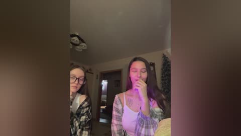A video of two young women with glasses and long hair in a dimly lit room. One is wearing a plaid shirt, the other a white top, both looking at a mirror.