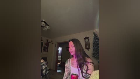 A video of a young woman with long dark hair in a casual outfit, sitting in a dimly lit living room with a ceiling fan, framed photos, and a Christmas tree.