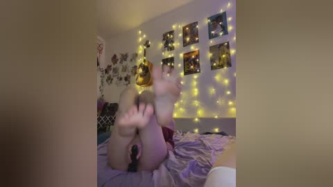 Video of a nude woman with fair skin and blonde hair, lying on a bed with purple sheets, surrounded by yellow fairy lights and framed posters, in a cozy bedroom setting.