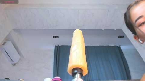 A video showing a person with short brown hair smiling, holding a bright orange dildo with a black base. The background features a grey marble wall with a black curtain and a white ceiling fan.