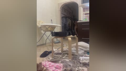 Video of a cluttered, dimly lit room with a small wooden chair, a metal ironing board, and scattered clothes on the floor. A doorway leads to a dark, archway.