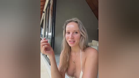 Media: Video of a blonde woman with fair skin, wearing a white bikini, standing in a sunlit attic room with wooden beams and large windows.