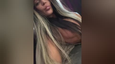 Media: A close-up video of a light-skinned woman with long, straight, blonde and dark brown hair, wearing a beige top, puckering her lips in a kiss. Background is blurred.