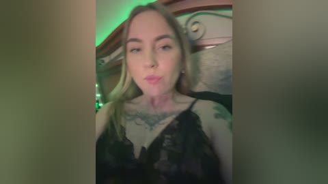 Media: A video of a young woman with pale skin and long blonde hair, wearing a black lace dress, sitting on a wooden chair with a floral-patterned cushion. The background has green lighting and decorative elements.