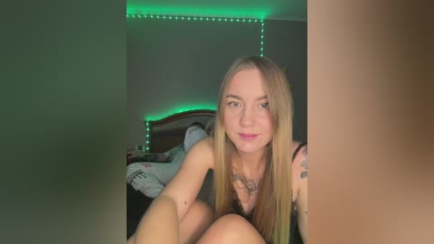 Media: A video of a young, fair-skinned woman with long blonde hair, wearing a black bra, sitting on a bed with a wooden headboard and green LED lights.