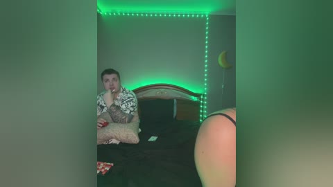 Media: A video of a young man with tattoos, sitting on a bed with a dark blanket, wearing a Hawaiian shirt, holding a red object, in a dimly lit room with green LED strip lighting.