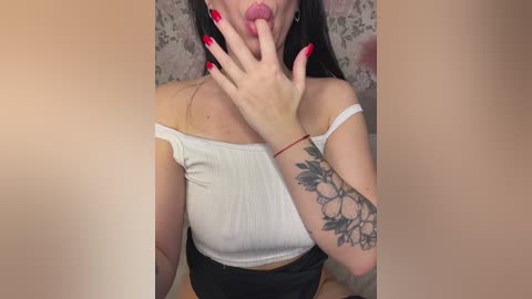 Video of a woman with long black hair, wearing a white off-shoulder crop top, red nails, and a floral tattoo on her arm, licking her lips seductively, against a floral-patterned background.