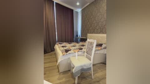 Media: Video of a cozy bedroom with a queen-sized bed covered in a quilted comforter, featuring a beige headboard, a white chair with a gray throw, and a wooden floor.