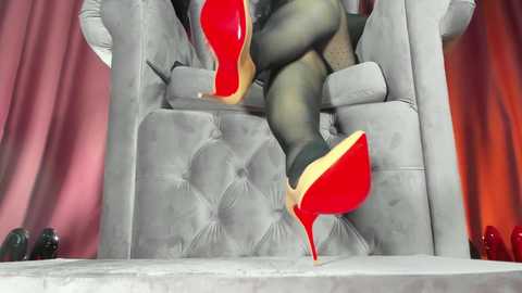 Video of a woman in black stockings and red high heels, sitting on a plush grey armchair, with a pink and red backdrop, creating a glamorous, luxurious atmosphere.