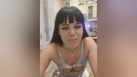 Video of a young woman with fair skin, long black hair, and bangs, wearing a revealing light blue top with cutouts, in a modern kitchen.