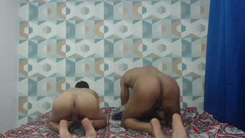 Video of two naked men with muscular builds, tan skin, and short dark hair, kneeling on a bed with a red, green, and white patterned blanket. They face each other, buttocks prominent. Background: patterned wallpaper and blue curtain.