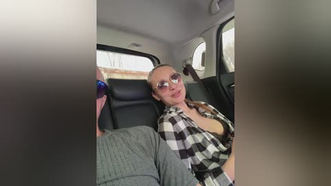 Video of a woman with light skin and glasses, wearing a black and white plaid shirt, reclining in a black leather car seat.