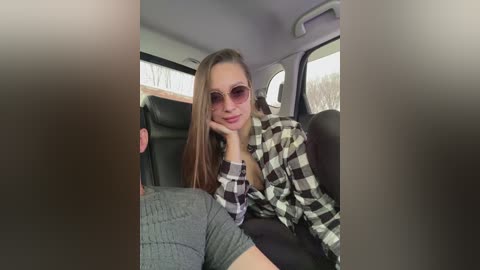 Video of a young woman with long brown hair, wearing a black-and-white plaid shirt and sunglasses, seated in a car.