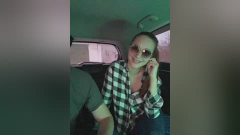 A video of a smiling young woman with light skin, fair hair tied back, wearing oversized sunglasses, a black and white checkered shirt, and jeans, seated in a dimly lit car.