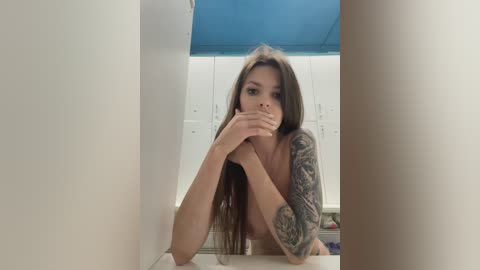 Media: Video of a topless young woman with long brown hair, medium-sized breasts, and a sleeve tattoo, covering her mouth, standing in a locker room with white lockers and blue ceiling.