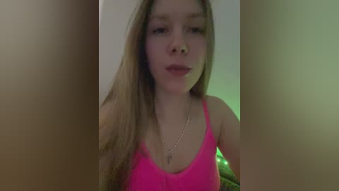 Video of a young, fair-skinned woman with long, straight brown hair, wearing a bright pink tank top, indoors under dim lighting, slightly out of focus.