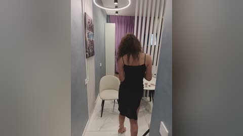 Media: Video of a woman in a black dress, standing in a narrow, modern hallway with white walls and a gray floor. She is facing away, with her long, curly brown hair cascading down.