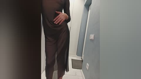 Media: Video of a woman in a sheer black dress, standing in a narrow hallway with light blue walls, white tile floor, and a white door. Her right hand rests on her hip.