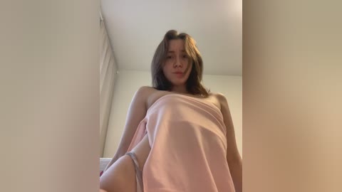 Media: A video of a young Asian woman with straight, shoulder-length brown hair, wearing a pink sheet, standing in a dimly lit room with beige walls.