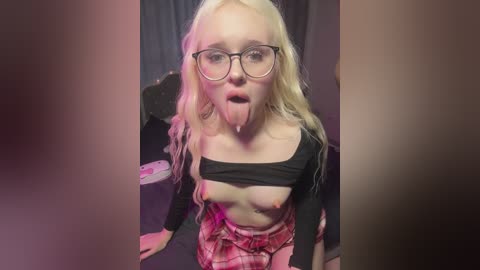 Media: Video of a blonde, fair-skinned woman with glasses, wearing a black crop top, exposing her small breasts, and a plaid skirt, licking her own cheek. Background is dark and blurry.