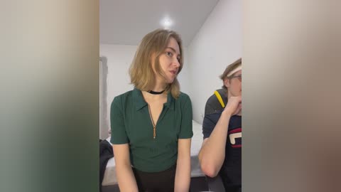 Video of a young Caucasian woman with shoulder-length blonde hair, wearing a dark green blouse and black choker, seated on a bed, talking to a man in a striped shirt.